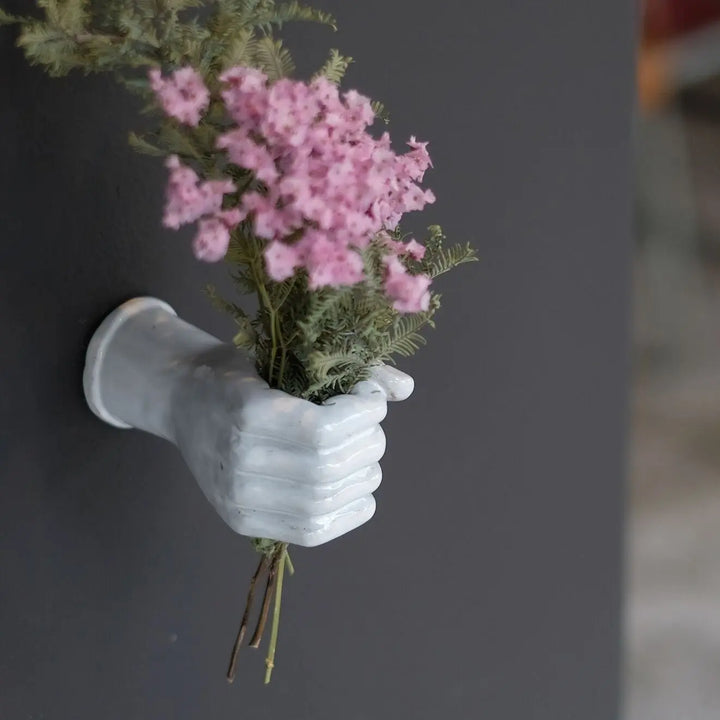 ピンクの花を持ち、手を握った形をした白いオブジェ。黒い壁に掛けられている。