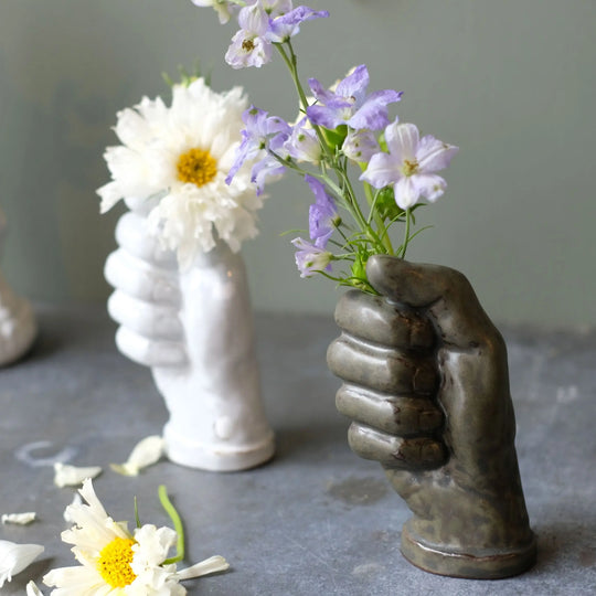 手が握る形をした花瓶。左奥に白い花が挿してある白、右手前に紫の花が挿してあるラスティ―グリーンの花瓶。