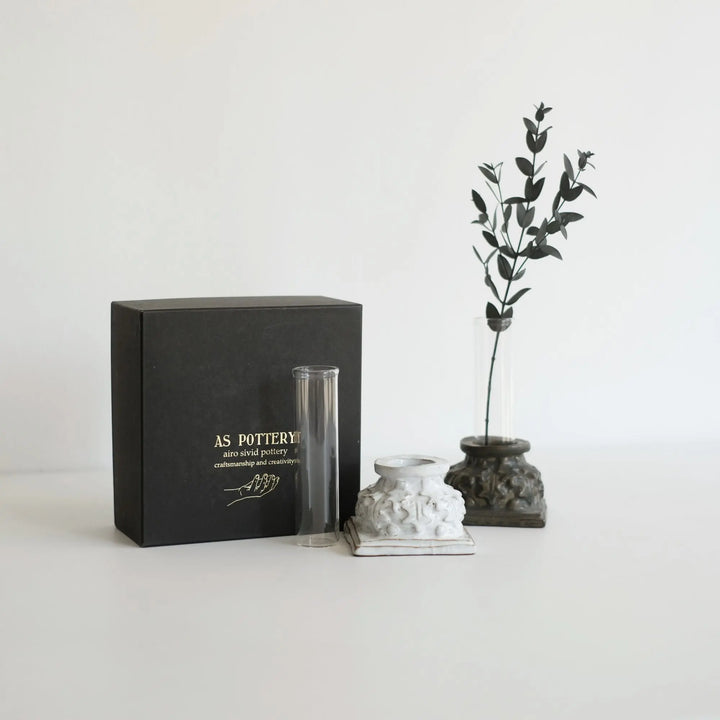 白い背景に、左から黒い商品用の箱、ガラスチューブ、白い陶器の台、植物が挿された青銅色の花瓶。